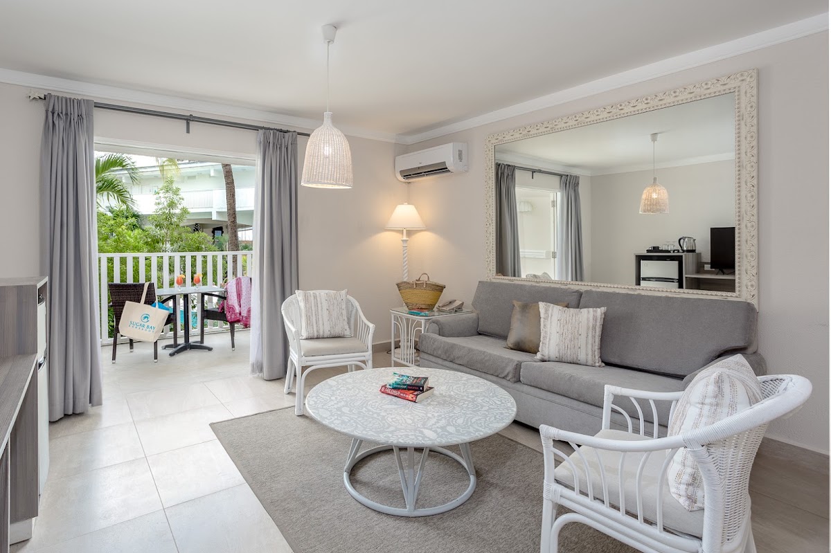 Spacious suite living room with gray sofa, wicker chairs, and round table opening to a balcony with palm trees at Sugar Bay Barbados.
