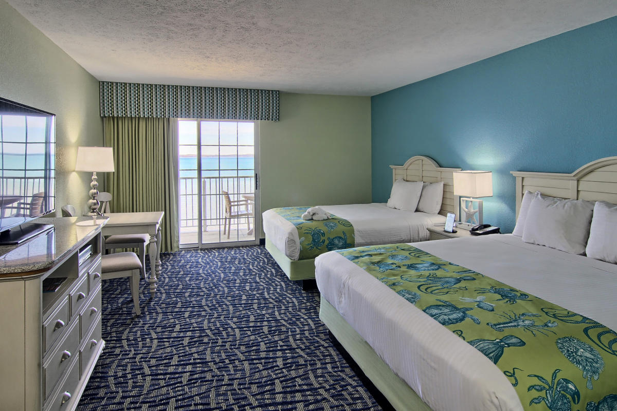 Resort room with two queen beds, ocean view balcony, green and blue walls, patterned carpet, and coastal scenery visible through glass doors.