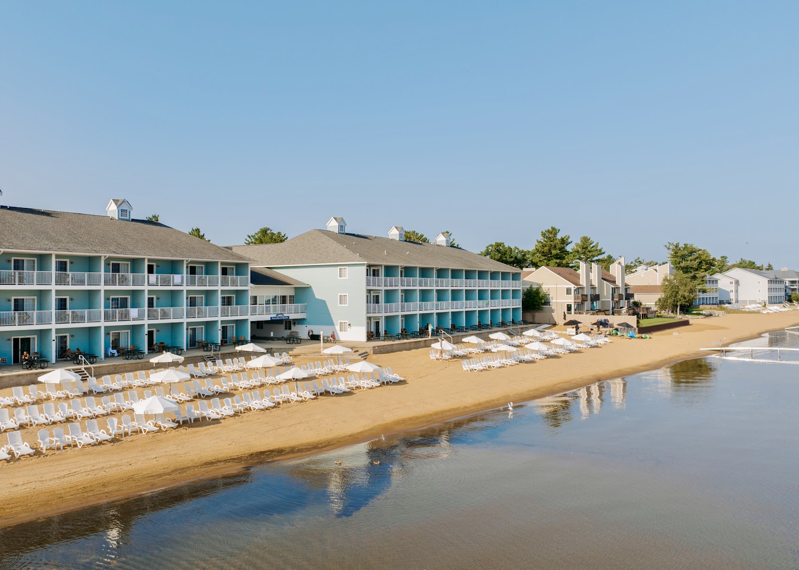 Turquoise and cream-colored resort buildings with white balconies line a sandy beach dotted with white lounge chairs facing calm bay waters.