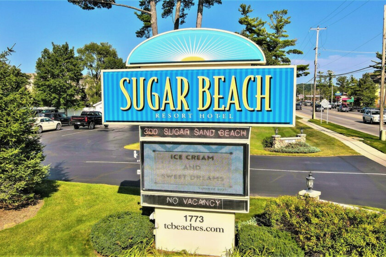 Blue and yellow Sugar Beach Resort Hotel entrance sign with turquoise sunburst design, parking lot and trees visible behind.