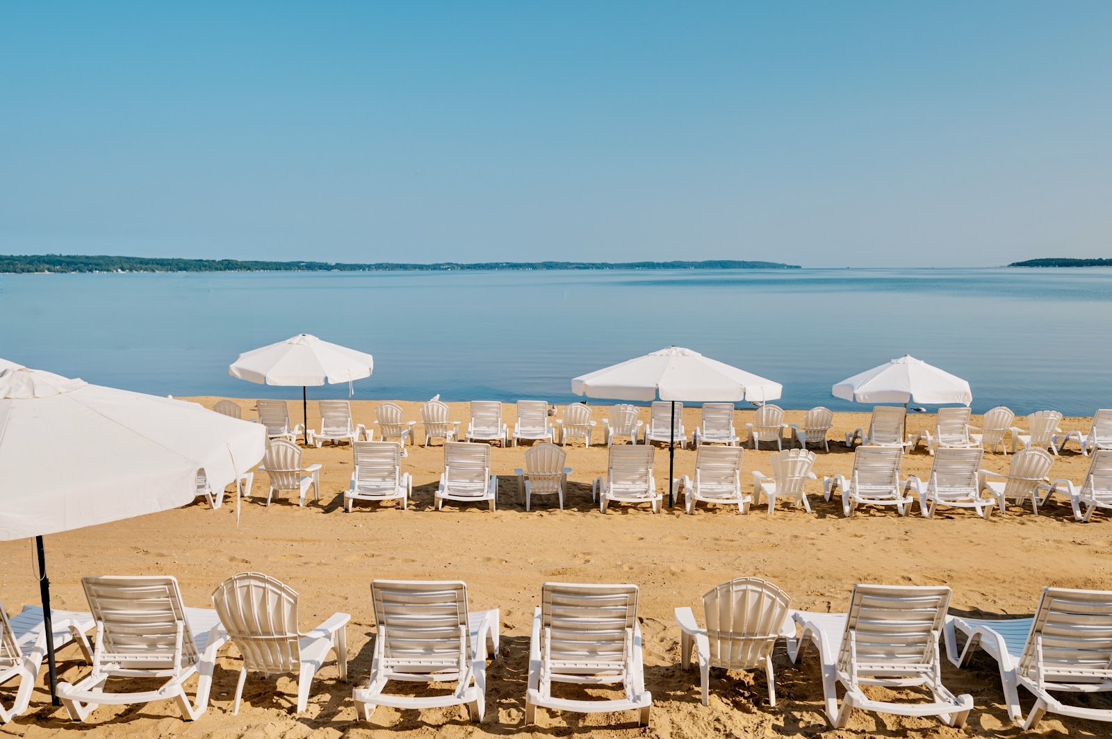 Sandy beach with rows of white lounge chairs and umbrellas facing calm turquoise water and distant shoreline.