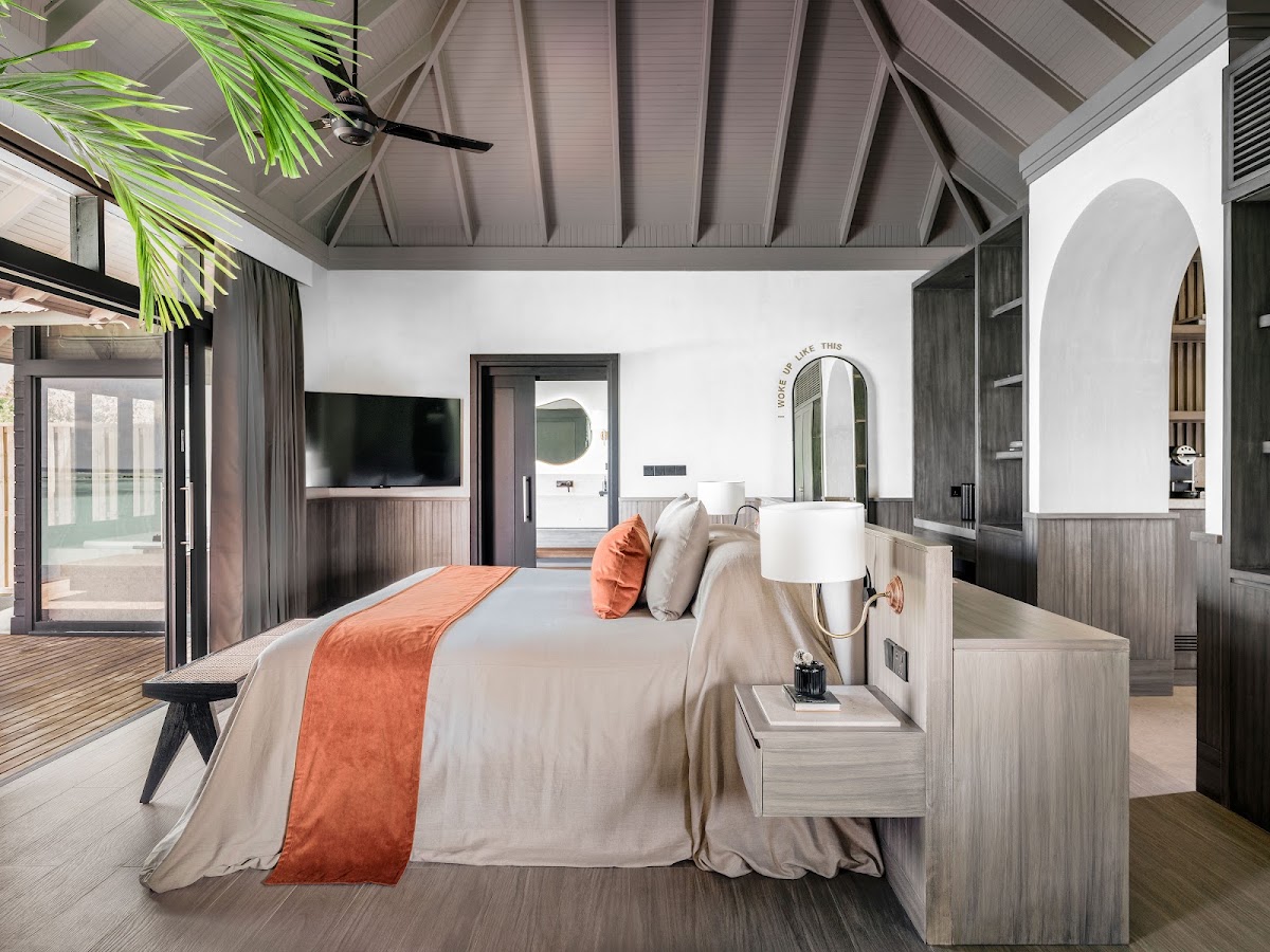 Modern resort villa bedroom with king bed, orange accent runner, wood nightstands, ceiling fan, TV, arched mirror, and glass doors opening to an ocean-view terrace.