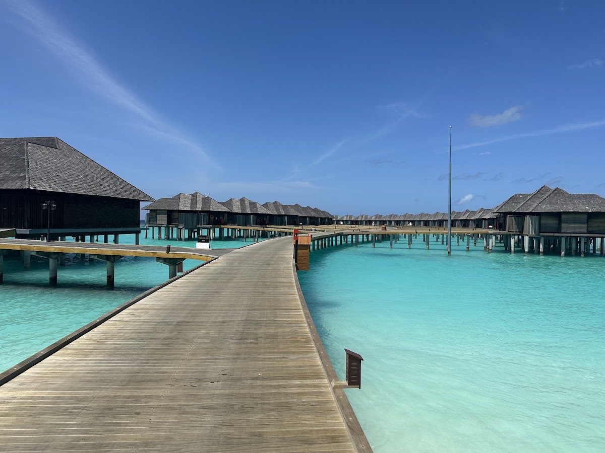 Wooden boardwalk lined with overwater bungalows at Sun Siyam Iru Fushi, surrounded by turquoise lagoon water under clear blue sky.