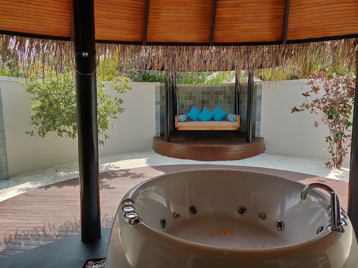 Overwater villa with sunken soaking tub in foreground, thatched roof shelter, and blue cushioned daybed on wooden deck surrounded by white walls and tropical plants.