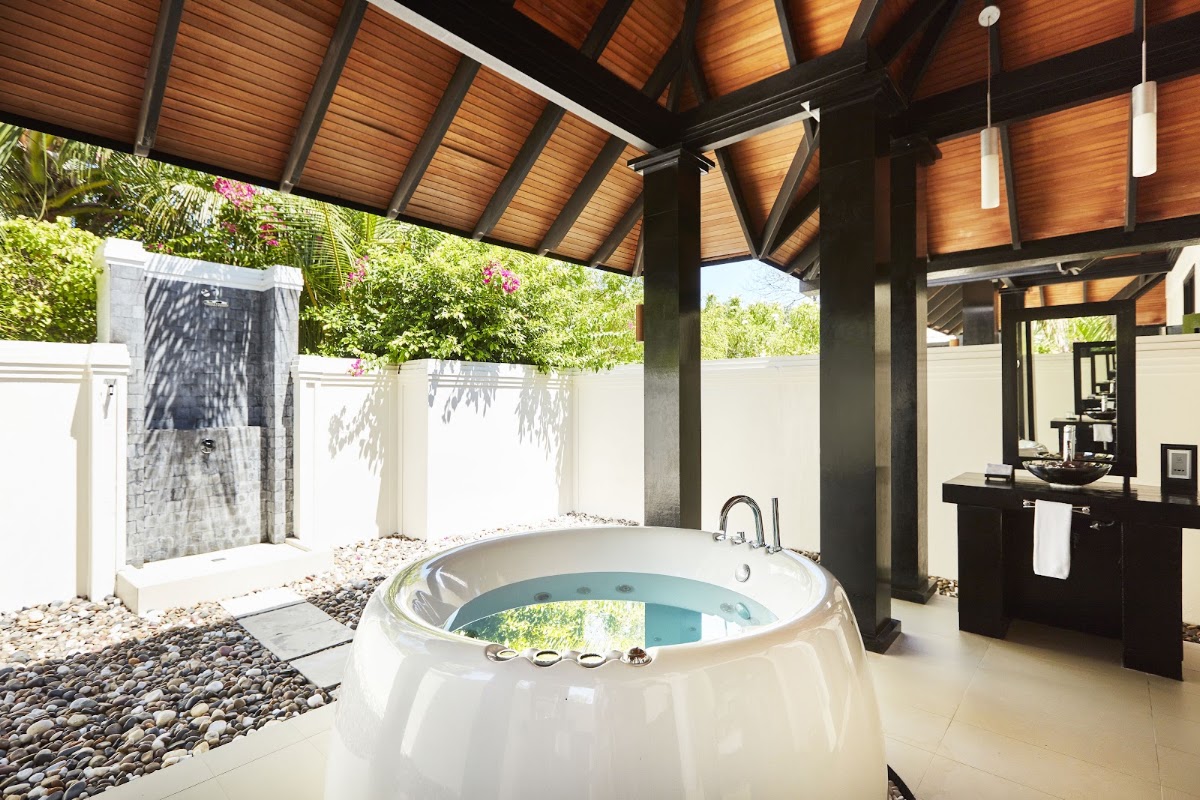 Outdoor bathroom with round soaking tub under wooden pergola, outdoor shower, and vanity at Sun Siyam Iru Fushi.