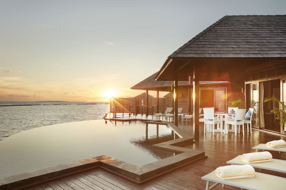Overwater bungalows at Sun Siyam Iru Fushi with wooden decks, white loungers, and dining furniture at sunset overlooking calm turquoise lagoon.