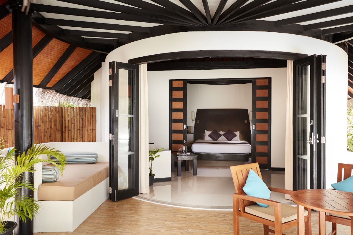 Sun Siyam Iru Fushi bungalow interior with open-air deck, four-poster bed visible through black wooden doors, and lounge seating.