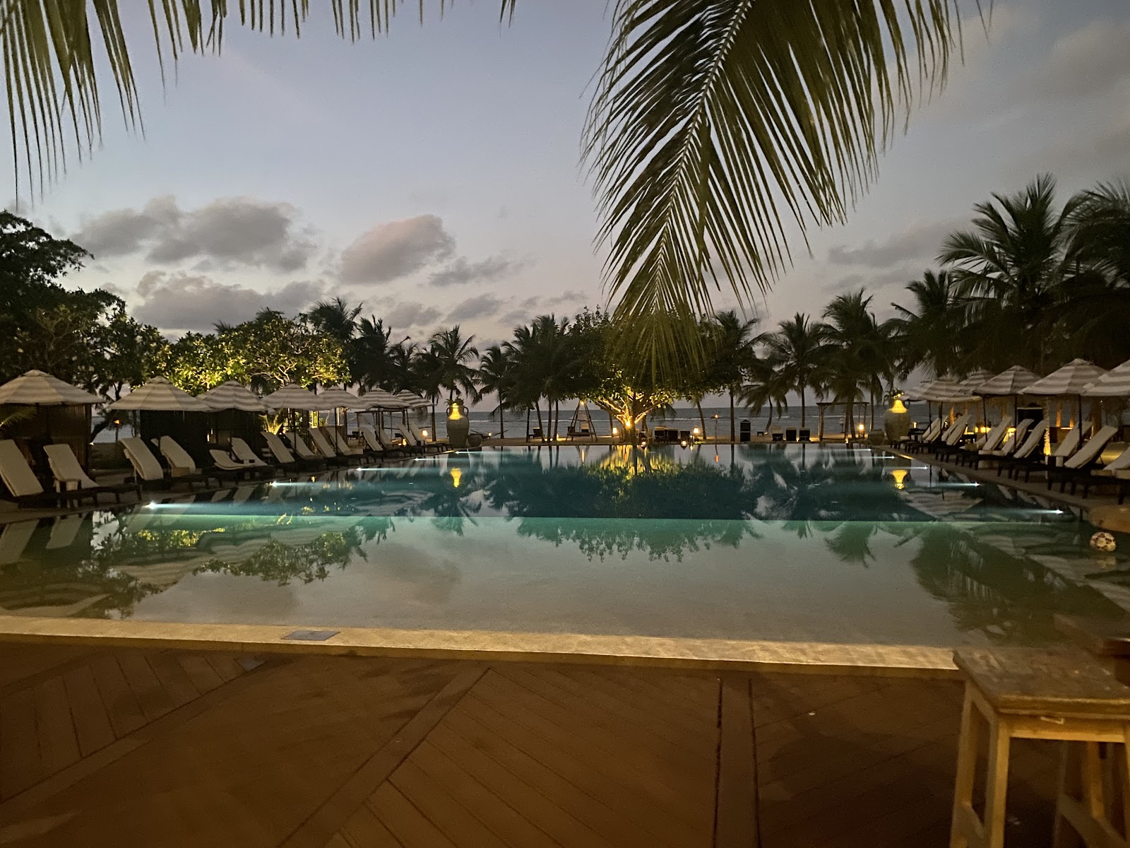 Twilight view of Sun Siyam Pasikudah's illuminated pool surrounded by lounge chairs, thatched umbrellas, and palm trees with ocean beyond.