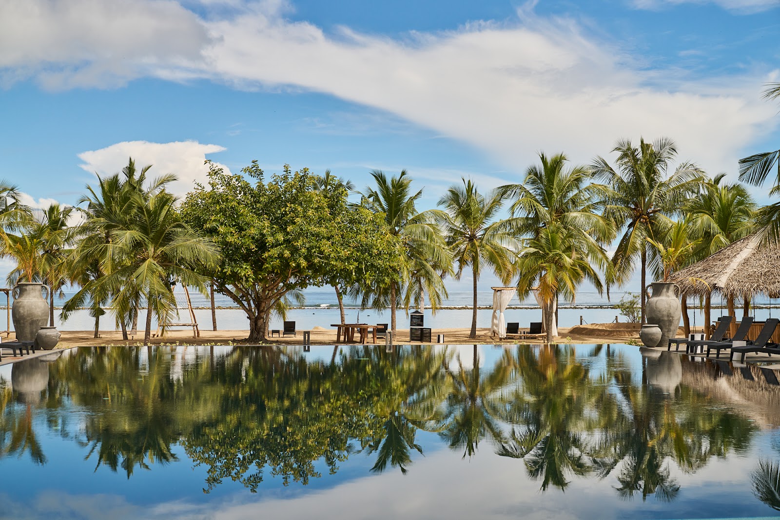 Infinity pool reflecting palm trees and ocean at Sun Siyam Pasikudah, with beach and thatched structures beyond.