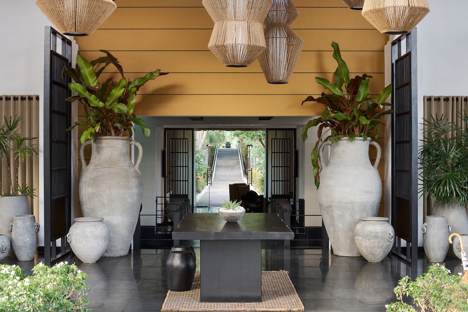 Covered entryway with gold ceiling, woven pendant lights, black doors, large white ceramic urns with plants, and black table overlooking gardens at Sun Siyam Pasikudah.