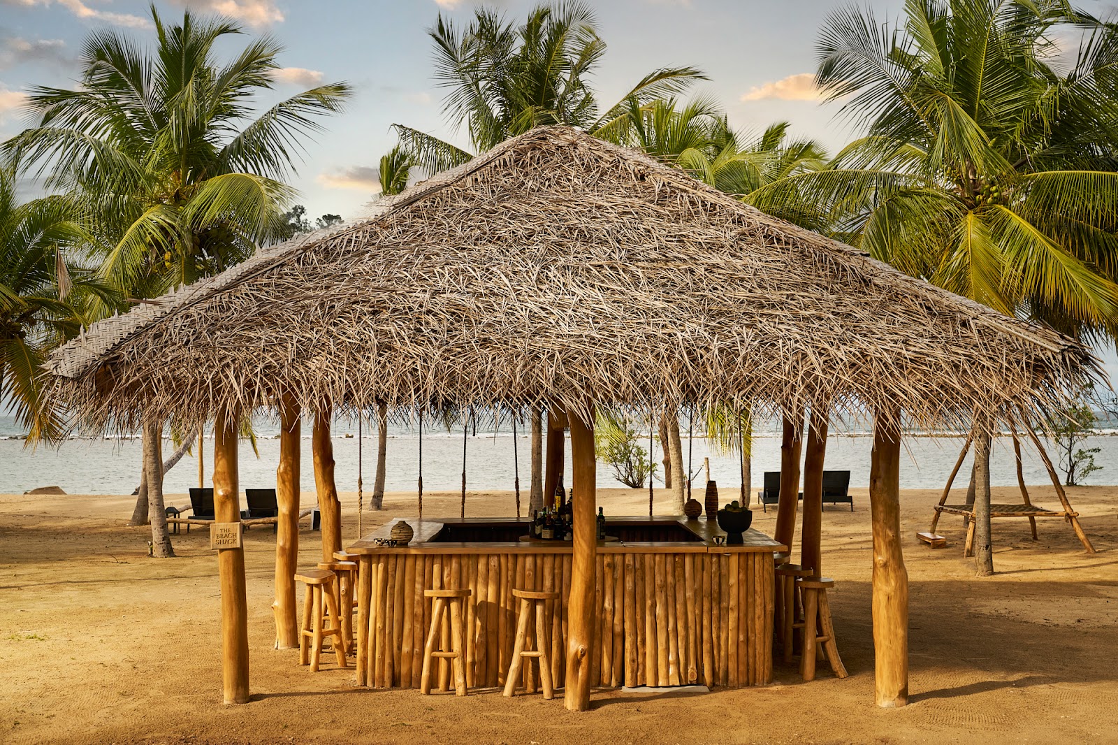 Thatched-roof bar with wooden counter and stools on sandy beach at Sun Siyam Pasikudah, surrounded by palm trees and water beyond.