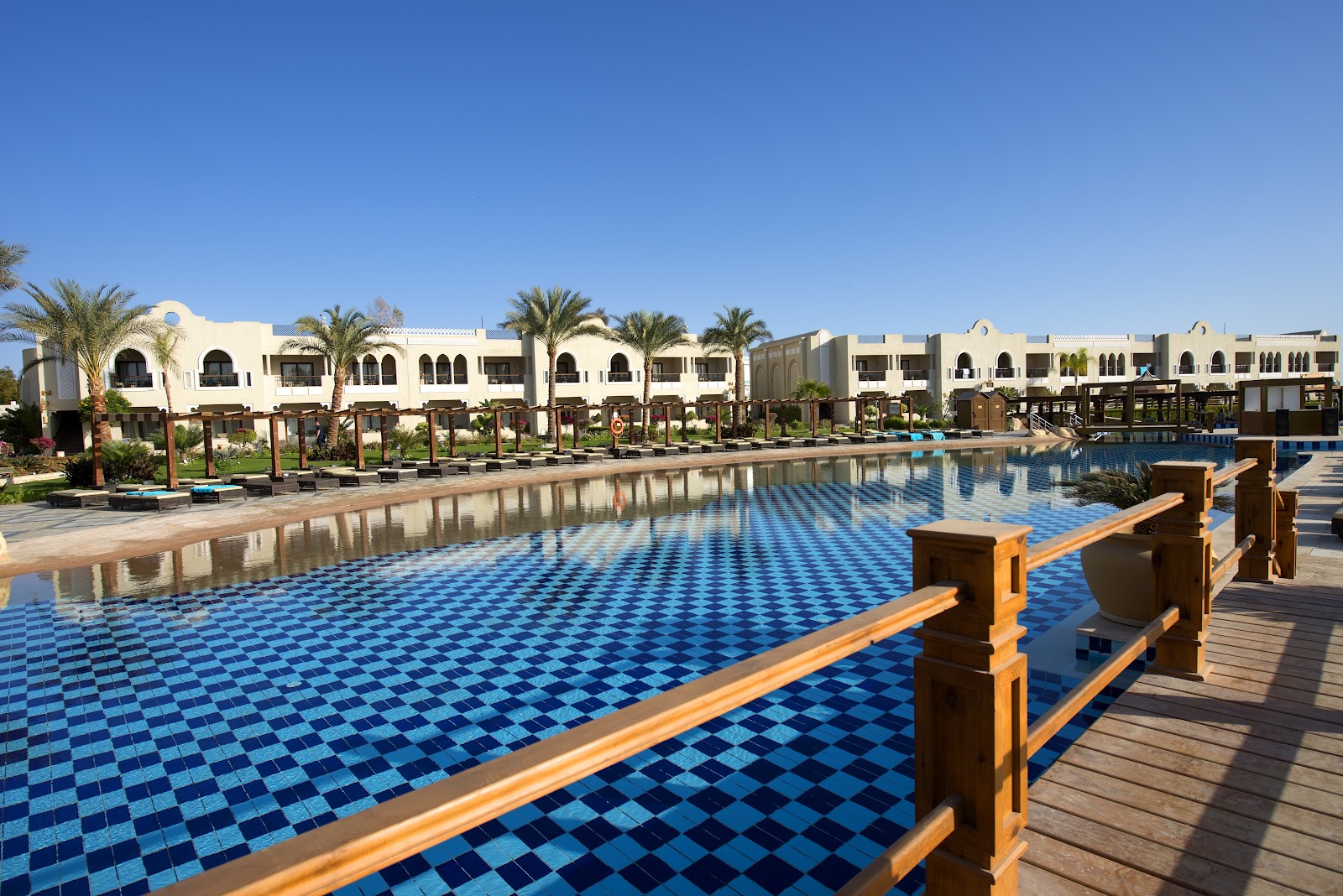 Blue tiled pool with wooden railings, palm trees, and white North African-style resort buildings under clear sky.