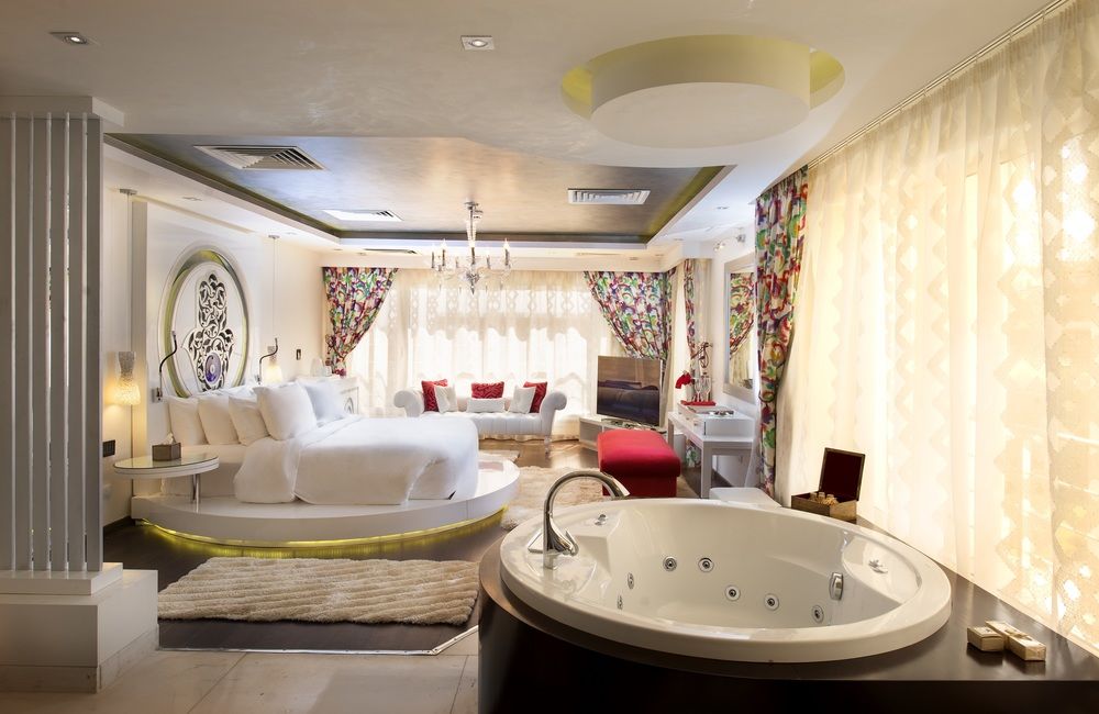 Luxurious suite bathroom with circular jetted tub in foreground, opening to bedroom with round bed, seating area, and floral curtains.