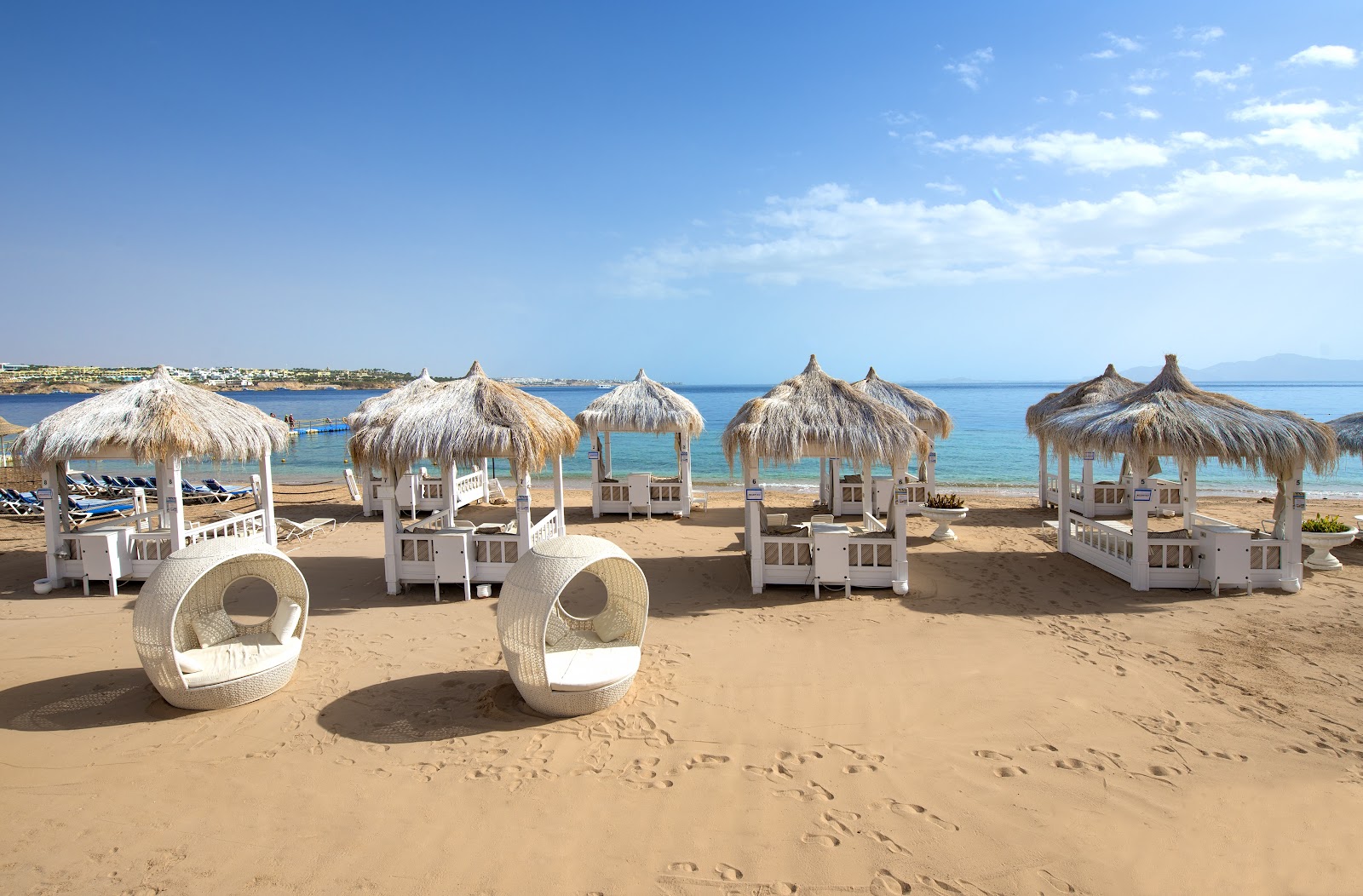 White woven lounge pods and thatched umbrellas line a sandy beach with turquoise water and distant coastline at Sunrise Arabian Beach Resort.