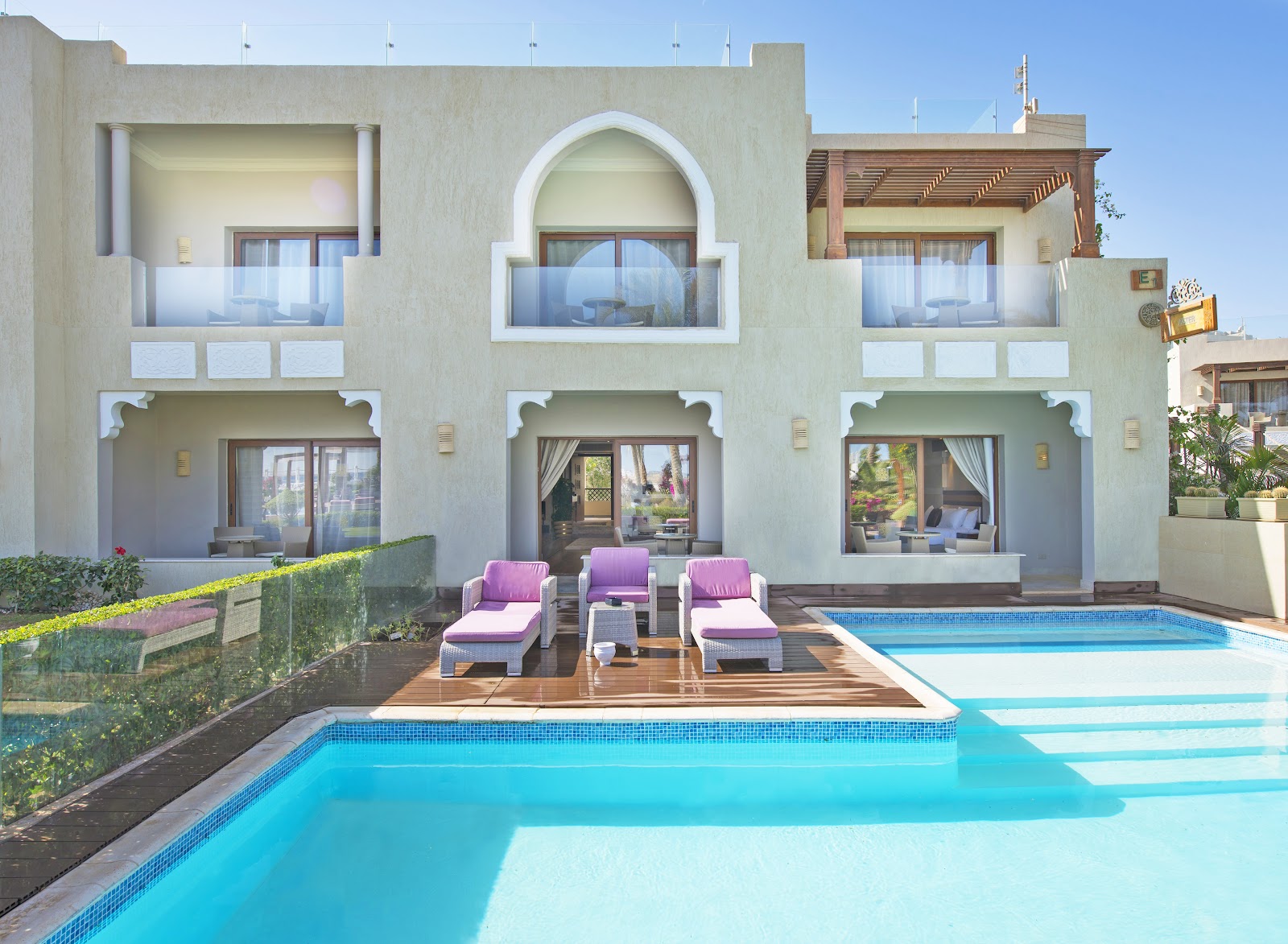 Pale green resort building with Moroccan-style arches overlooks two pools and wooden deck lined with pink lounge chairs.