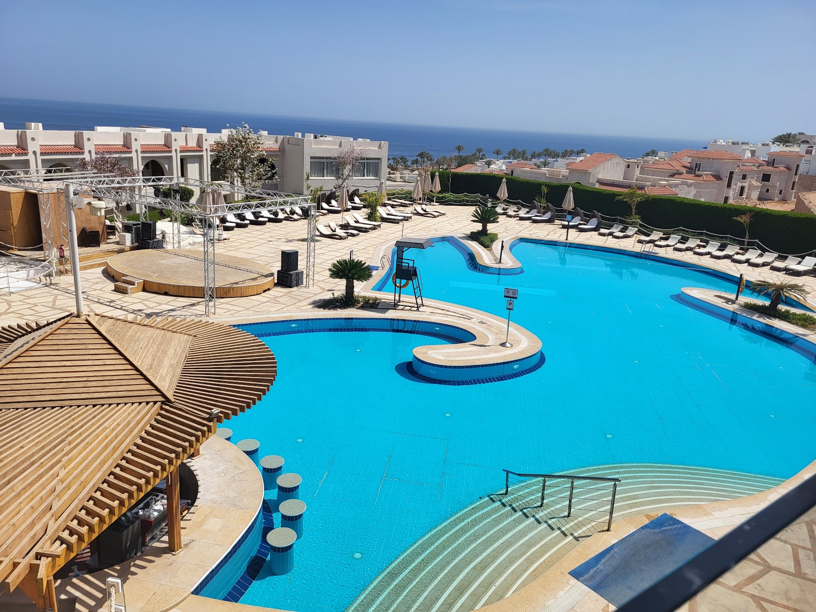 Bright blue pools with curved sections and in-pool seating, surrounded by sun loungers, wooden shade structures, and Mediterranean-style resort buildings overlooking the sea.