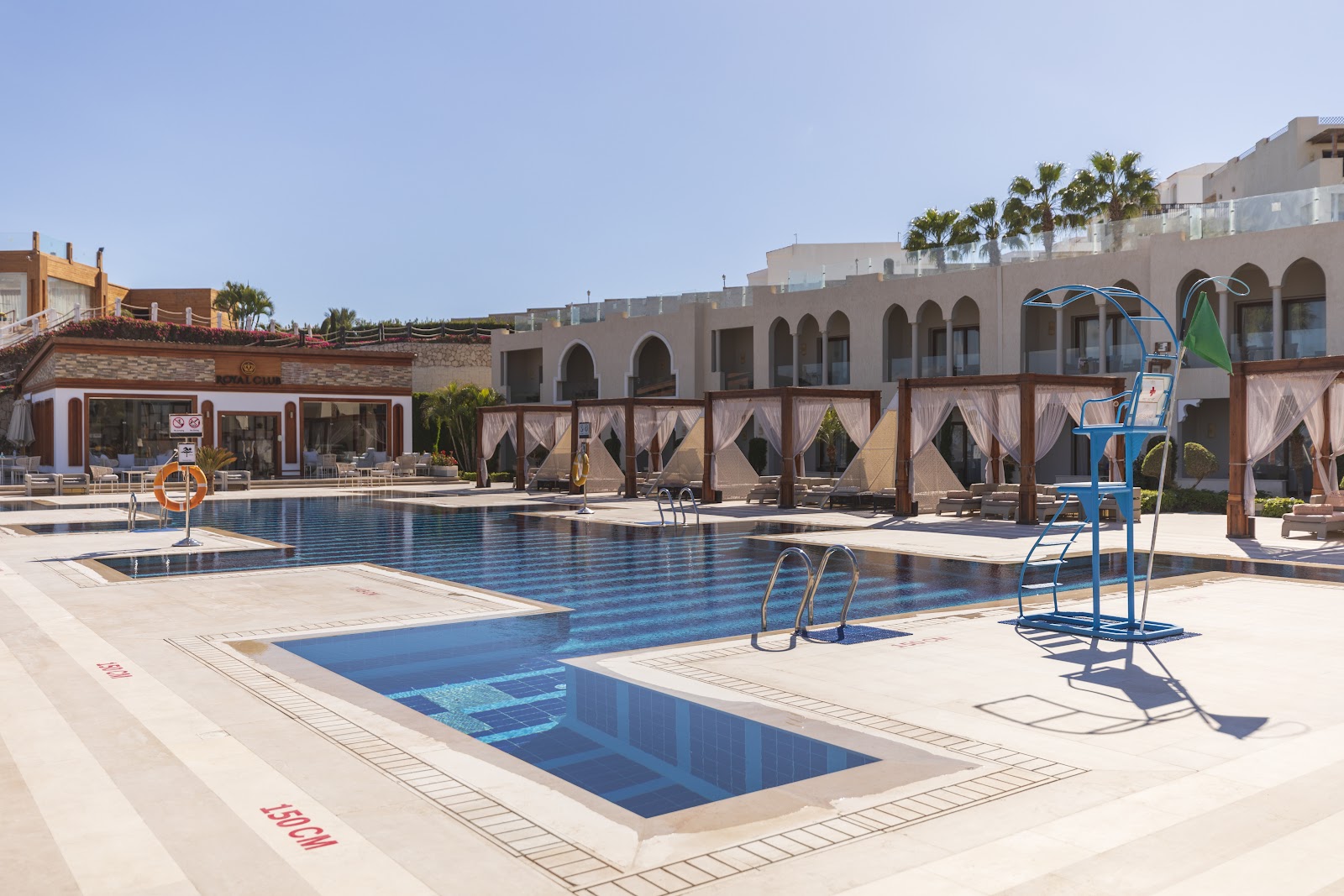 Resort pool area with multiple swimming pools, blue lifeguard chairs, tan cabanas with curtains, and arched architectural buildings under clear sky.