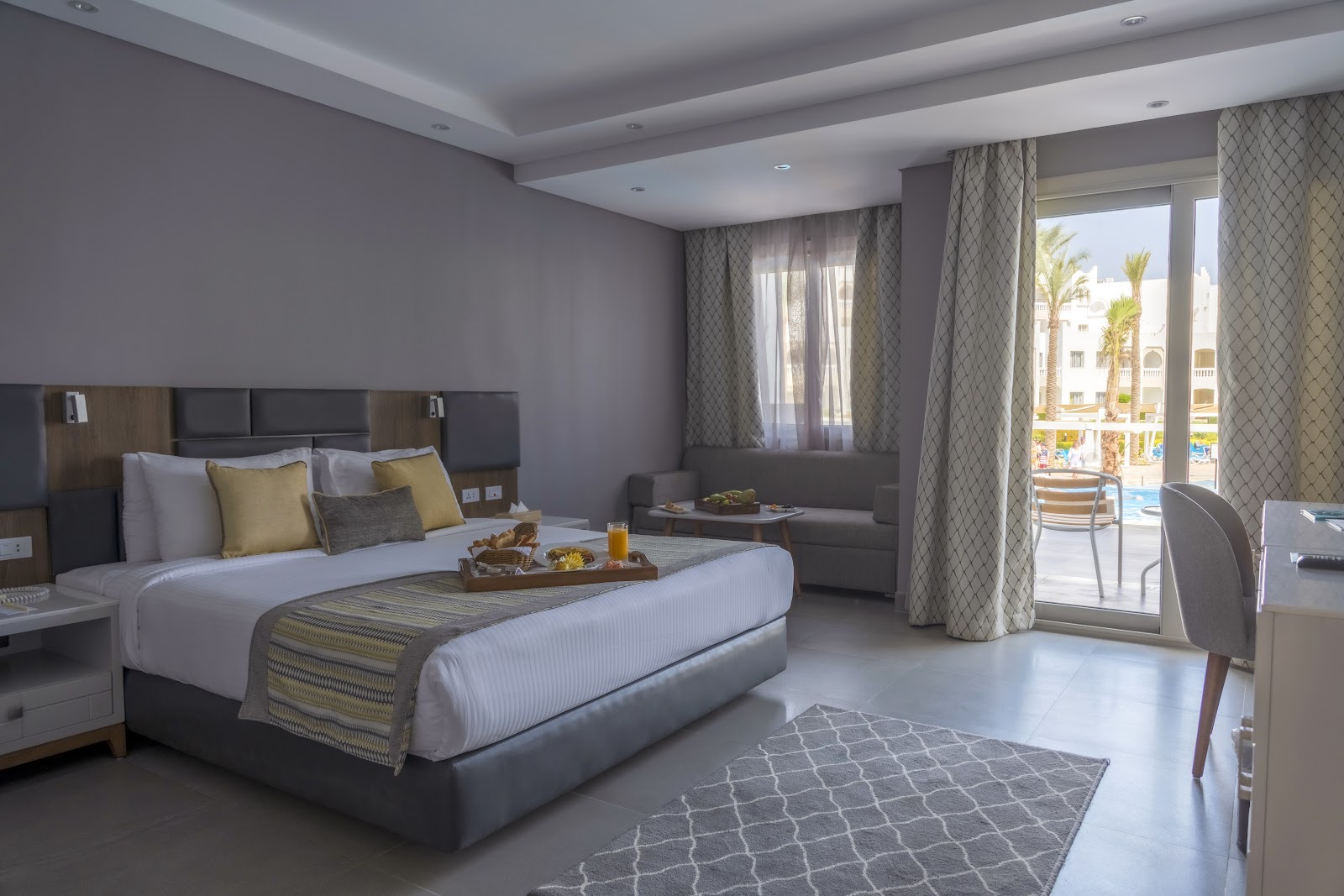 Modern hotel room with king bed, gray accent walls, patterned curtains, and balcony overlooking pool and palm trees at Sunrise Select Diamond Beach Resort.