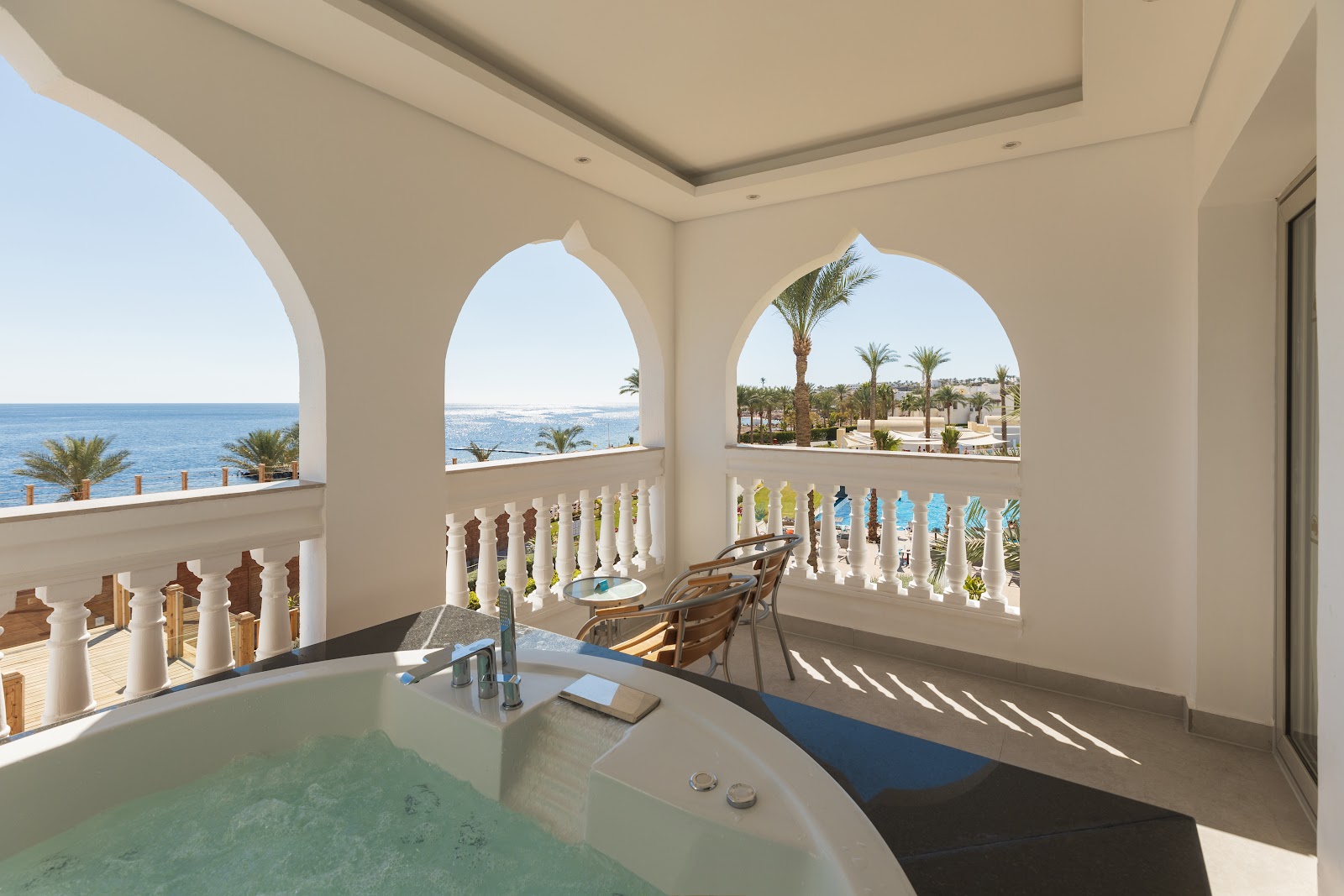 Bathroom with soaking tub and arched windows overlooking the sea, palm trees, and Sunrise Select Diamond Beach Resort grounds.