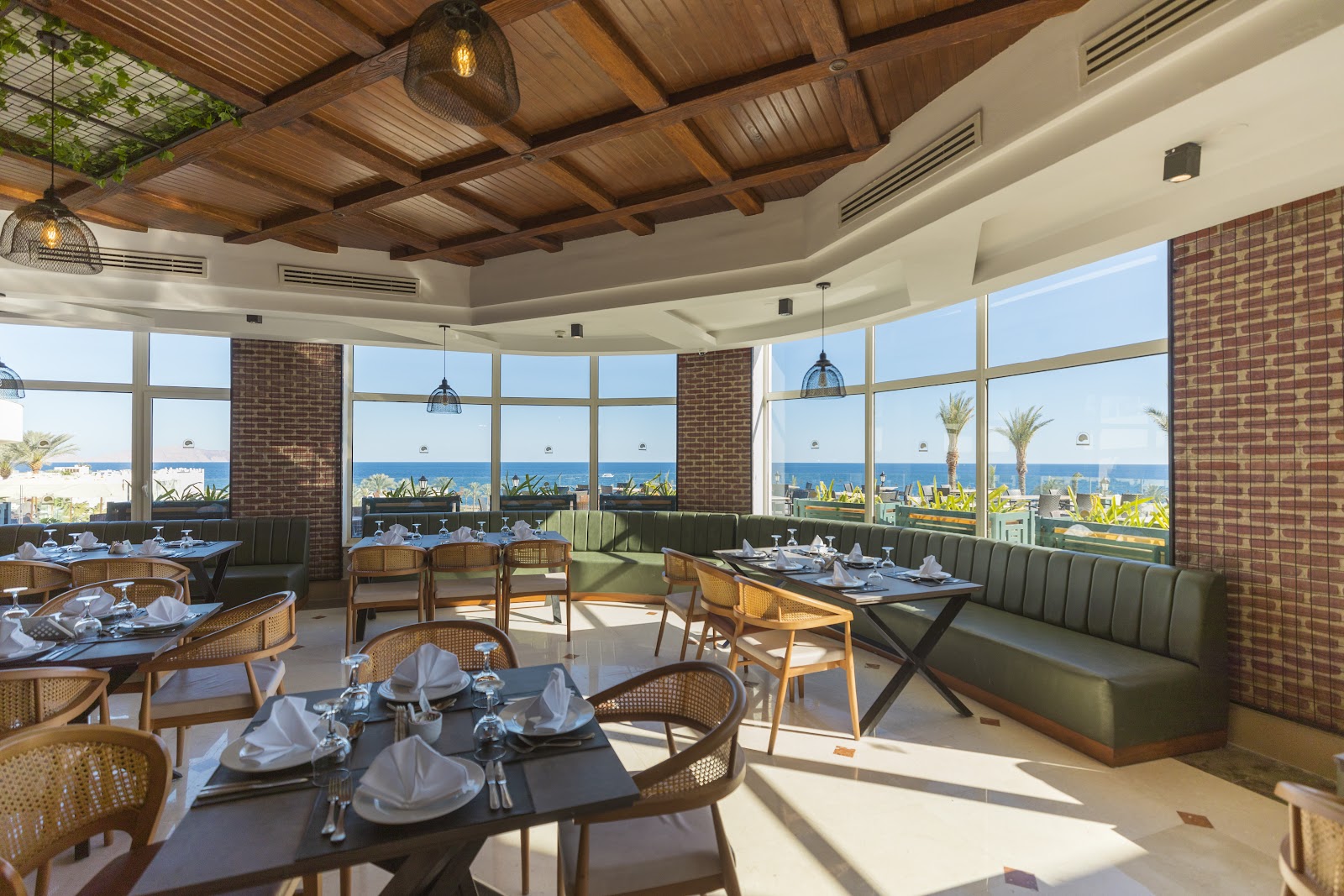 Dining room with set tables, rattan chairs, brick pillars, wood ceiling, and floor-to-ceiling windows overlooking the sea and palm trees.