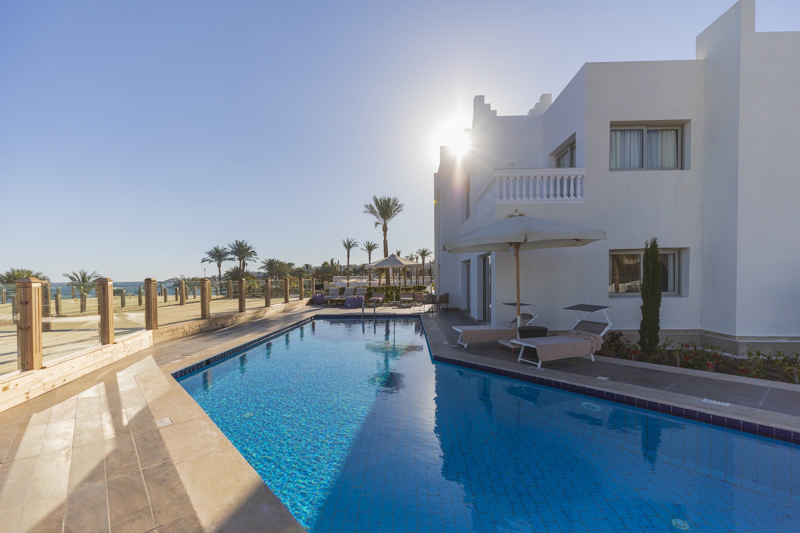 White resort building with a blue pool, sun loungers, palm trees, and beach beyond a wooden fence under clear sky.