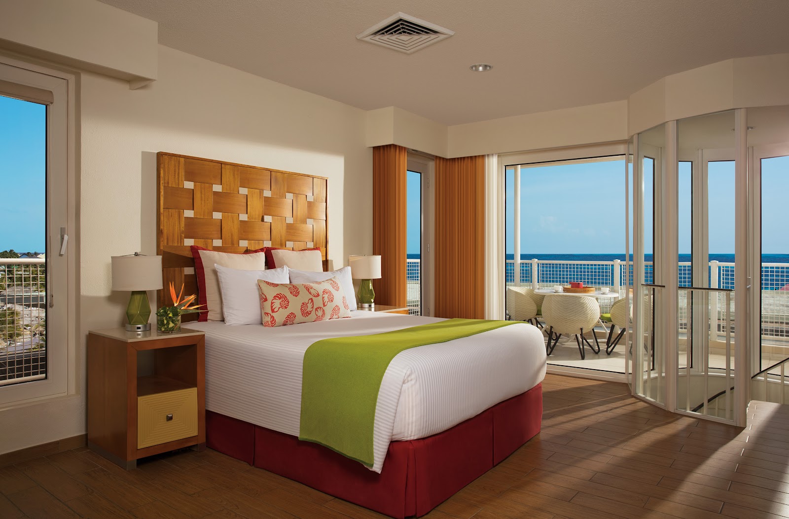 Resort guestroom with ocean-view balcony, queen bed with wooden headboard, and floor-to-ceiling windows overlooking blue water and horizon.