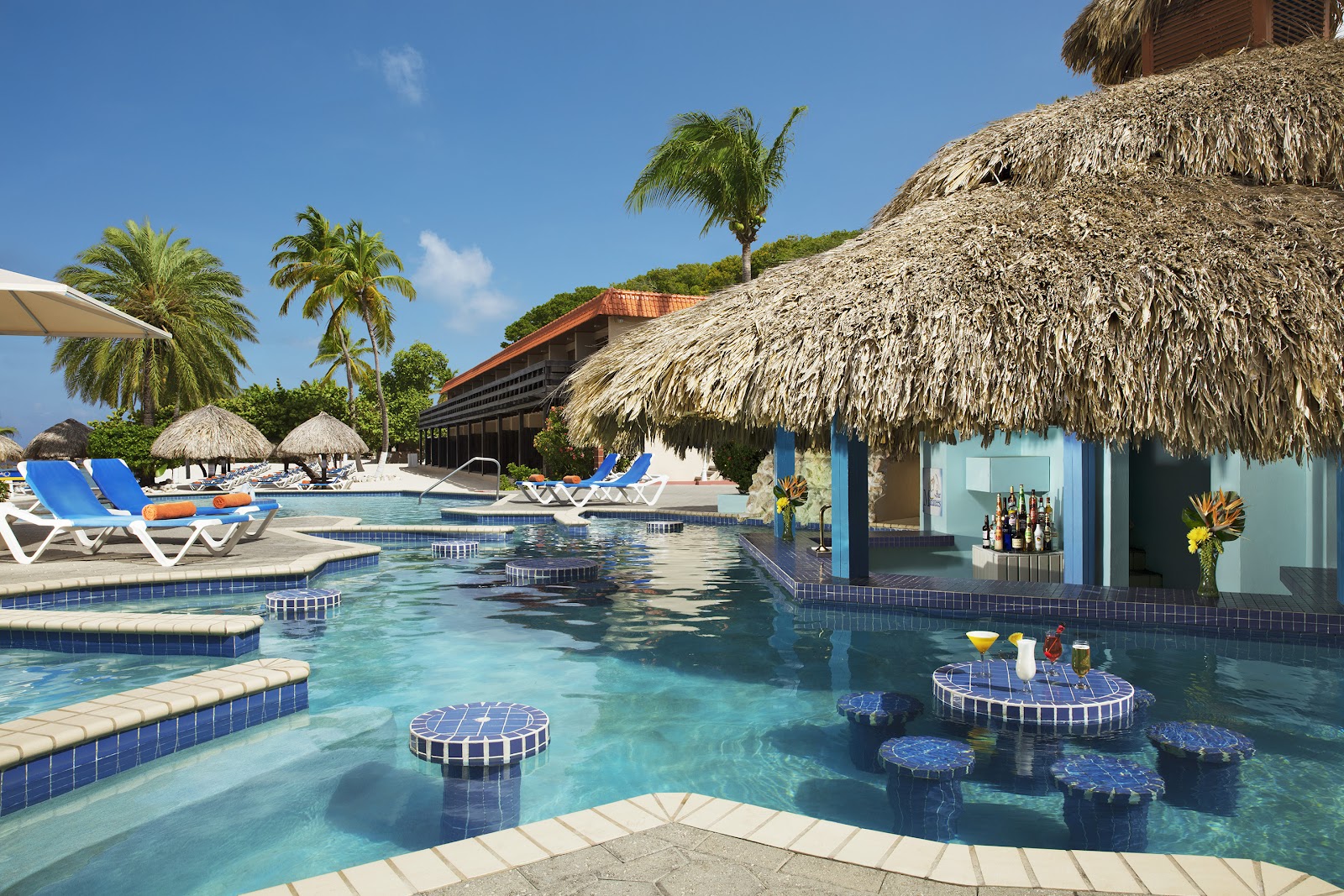 Sunscape Curaçao's pool area features turquoise water with floating bar platforms, thatched-roof cabanas, lounge chairs, and palm trees under clear blue sky.