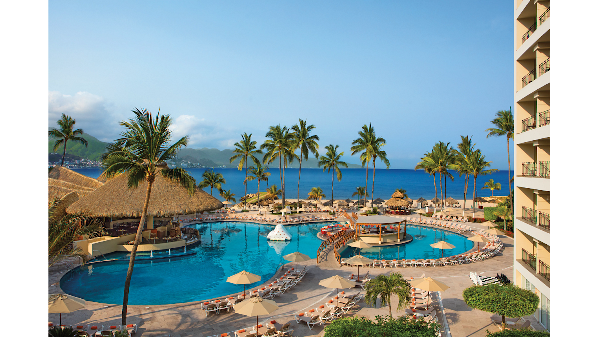 Curved pool with swim-up bar and fountains, surrounded by lounge chairs and umbrellas, with palm trees, beach, and mountains beyond at Sunscape Puerto Vallarta Resort & Spa.