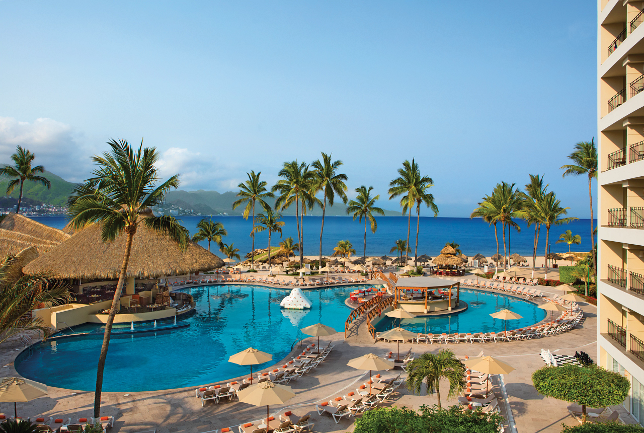 Sunscape Puerto Vallarta Resort & Spa pool area with palm trees, lounge chairs, umbrellas, and ocean view beyond the beach.