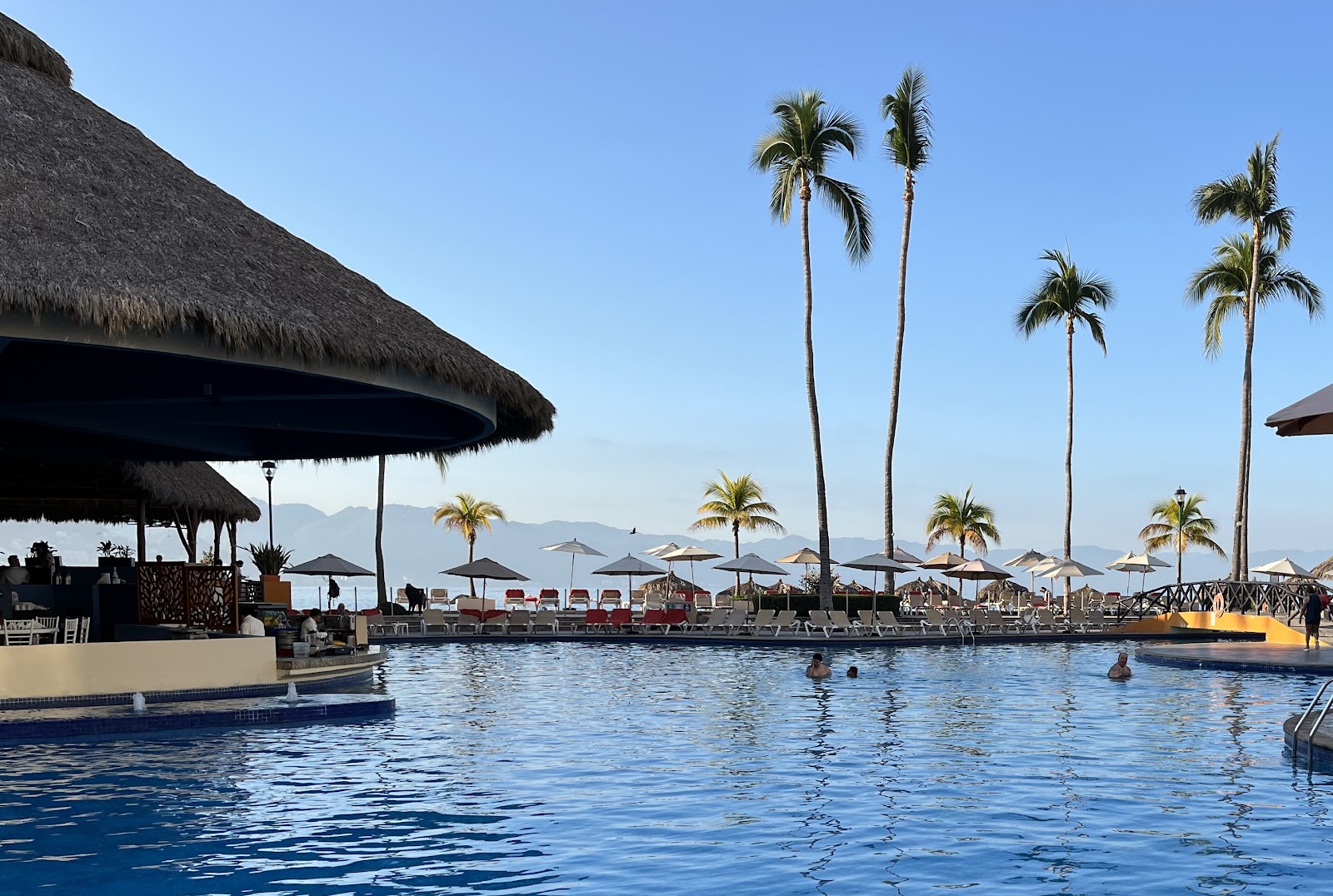 Curved pool at dusk with lounge chairs, thatched palapas, and tall palm trees, Sunscape Puerto Vallarta Resort & Spa visible in background.