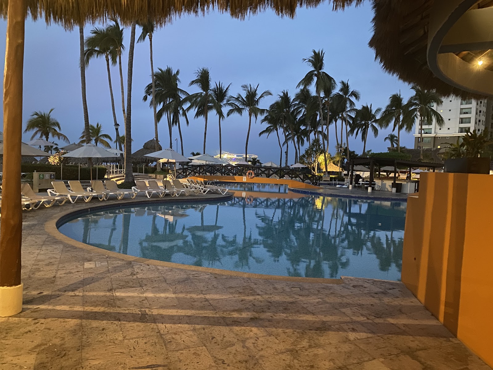 Sandy beach with thatched umbrellas, blue lounge chairs, and turquoise ocean backed by green mountains under clear sky.