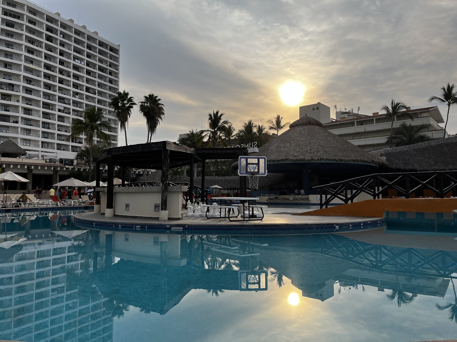 Turquoise pool with basketball hoop at sunset, palm trees and high-rise hotel building in background at Sunscape Puerto Vallarta.
