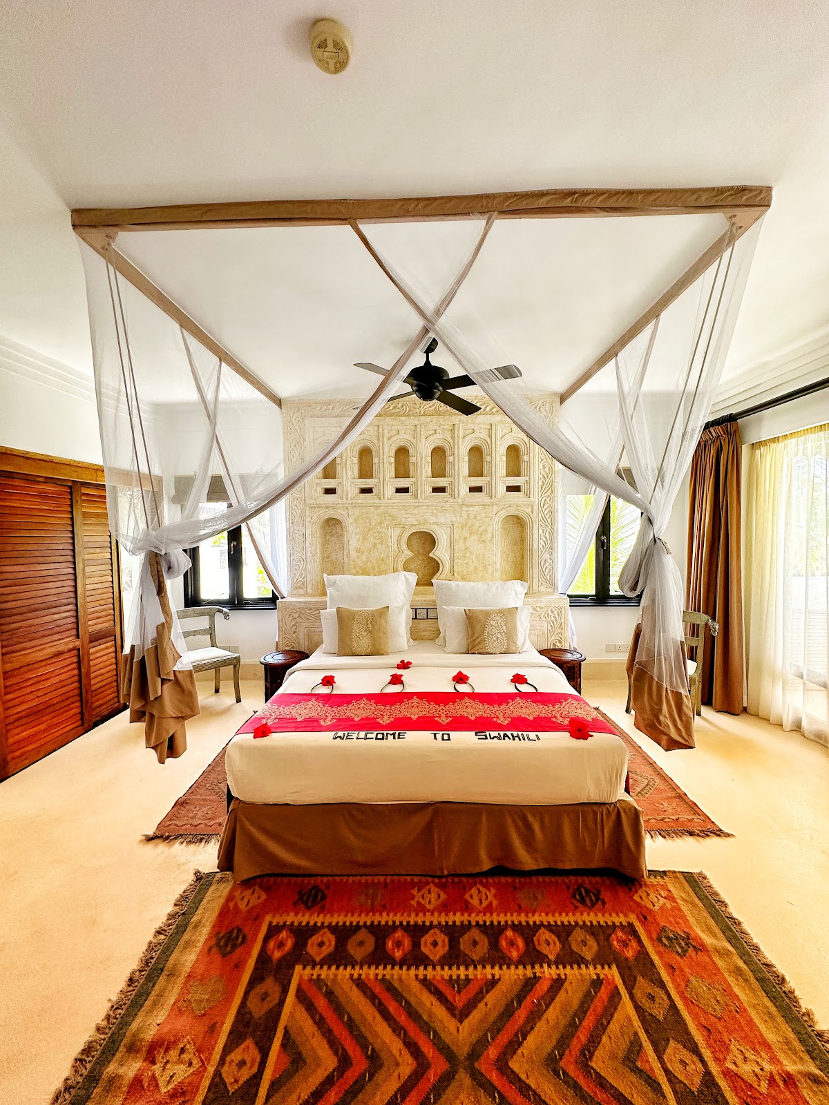 Bedroom suite with four-poster canopy bed draped in sheer white fabric, decorated with red rose petals and welcome banner, ornate carved headboard, and patterned area rug.