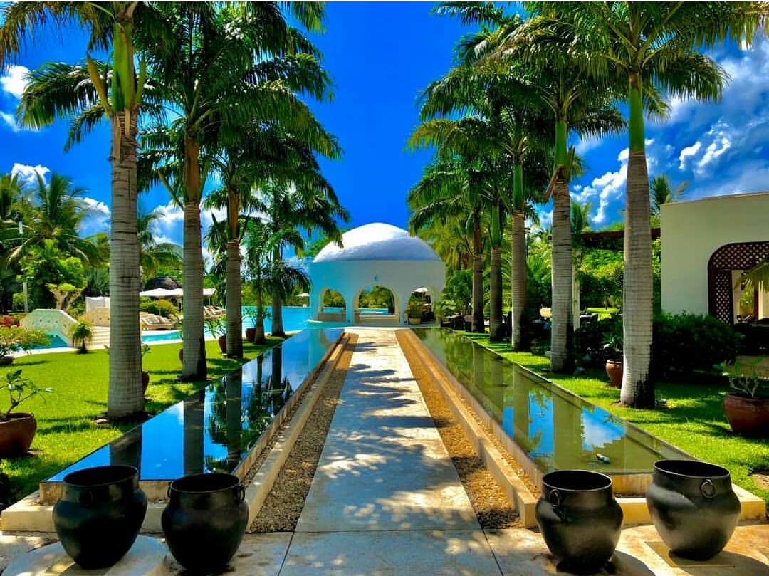 Palm-lined water channel with black ceramic pots leads to white domed structure at Swahili Beach Resort, flanked by green lawns and pool area under blue sky.