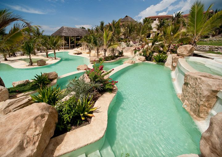 Turquoise lazy river winds through palm trees and rocky outcrops at Swahili Beach Resort, with thatched structures visible beyond.