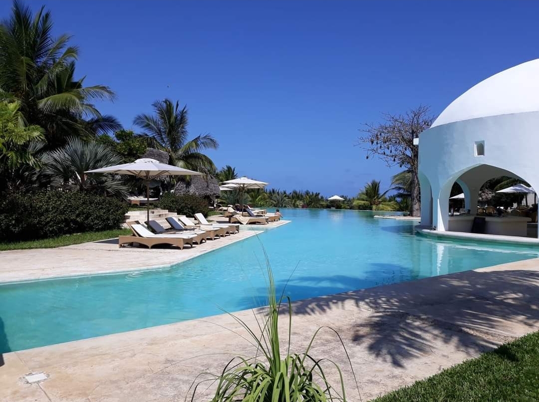 Turquoise pool with lounge chairs and white umbrellas at Swahili Beach Resort, palm trees and white arched structures along the water.