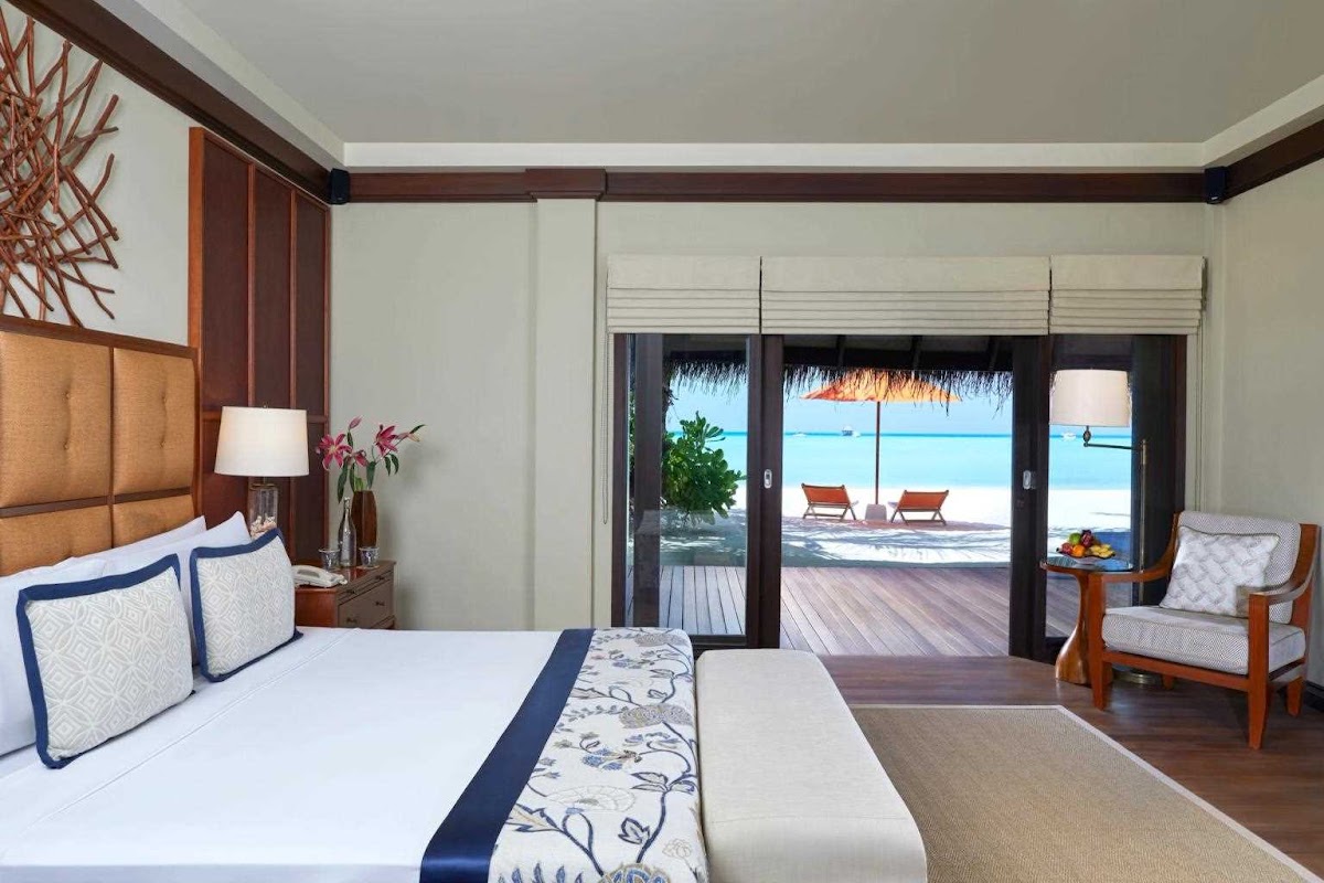 Beachfront resort bedroom with king bed, ocean view through glass doors, thatched roof shelter and lounge chairs visible on deck.