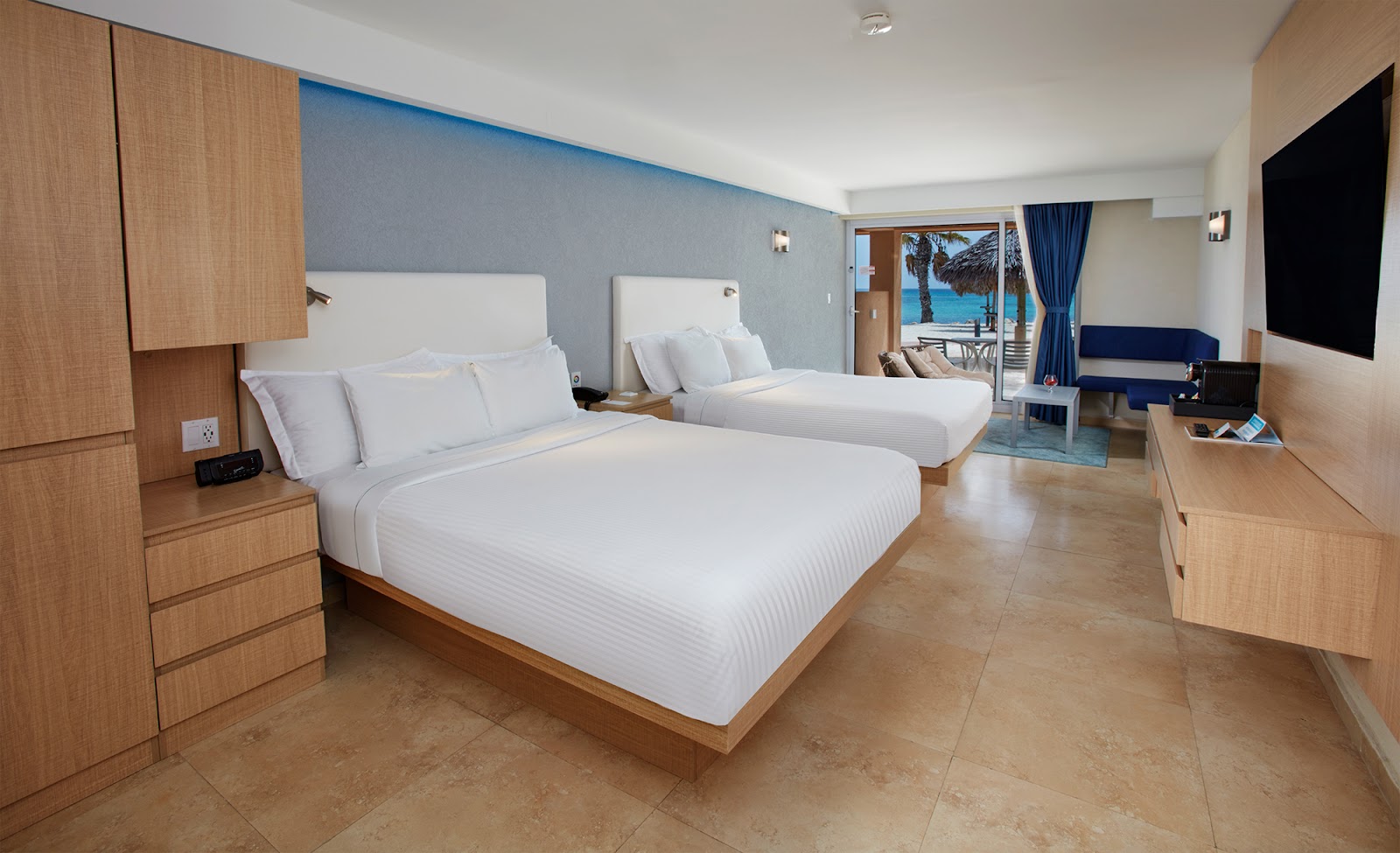 Resort guestroom with two queen beds, wood furnishings, blue accent wall, and ocean view through balcony doors.