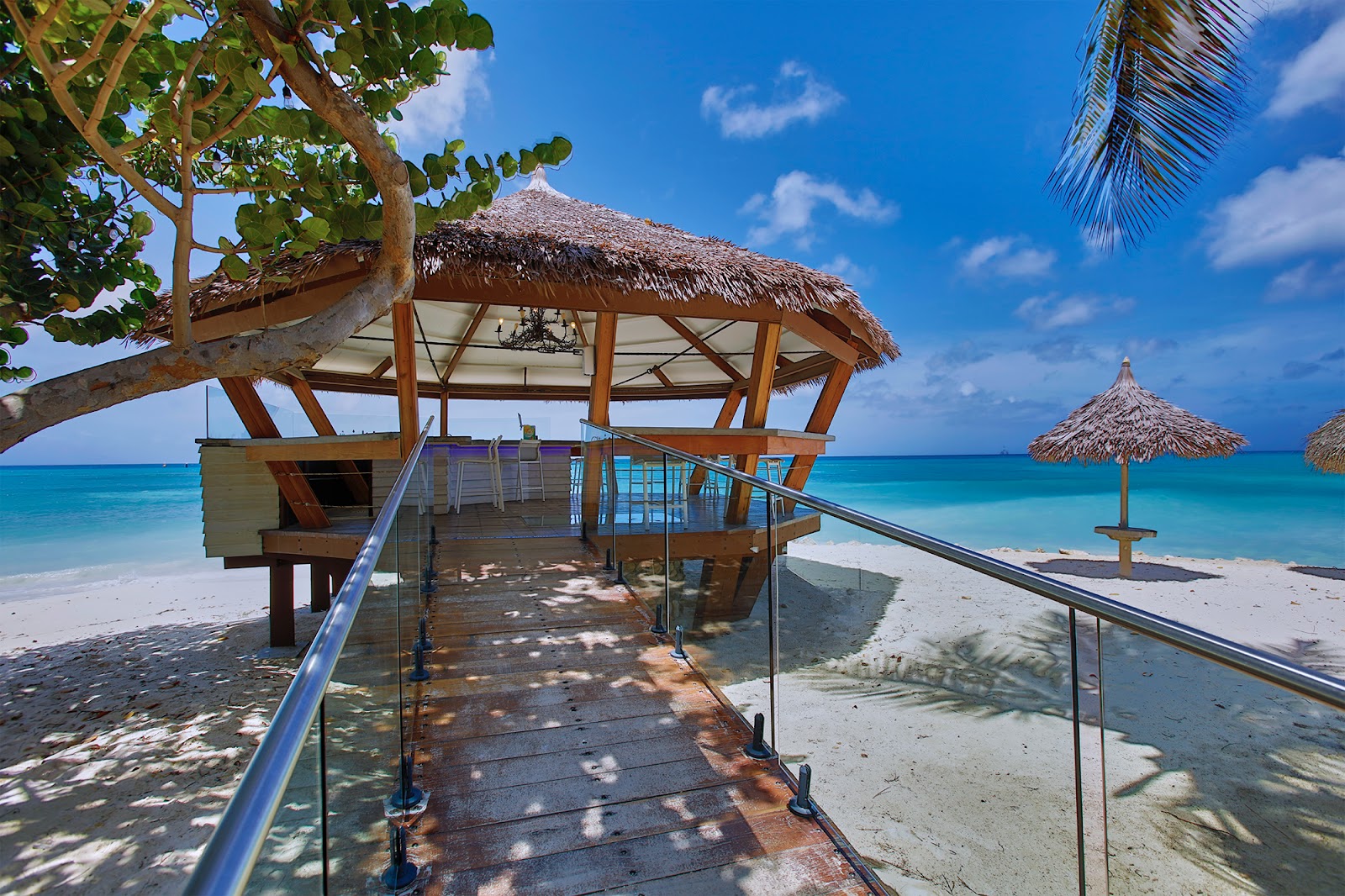 Wooden pier with thatched-roof pavilion extends into turquoise water at Tamarijn Aruba, with white sand beach and palm-framed umbrellas beyond.