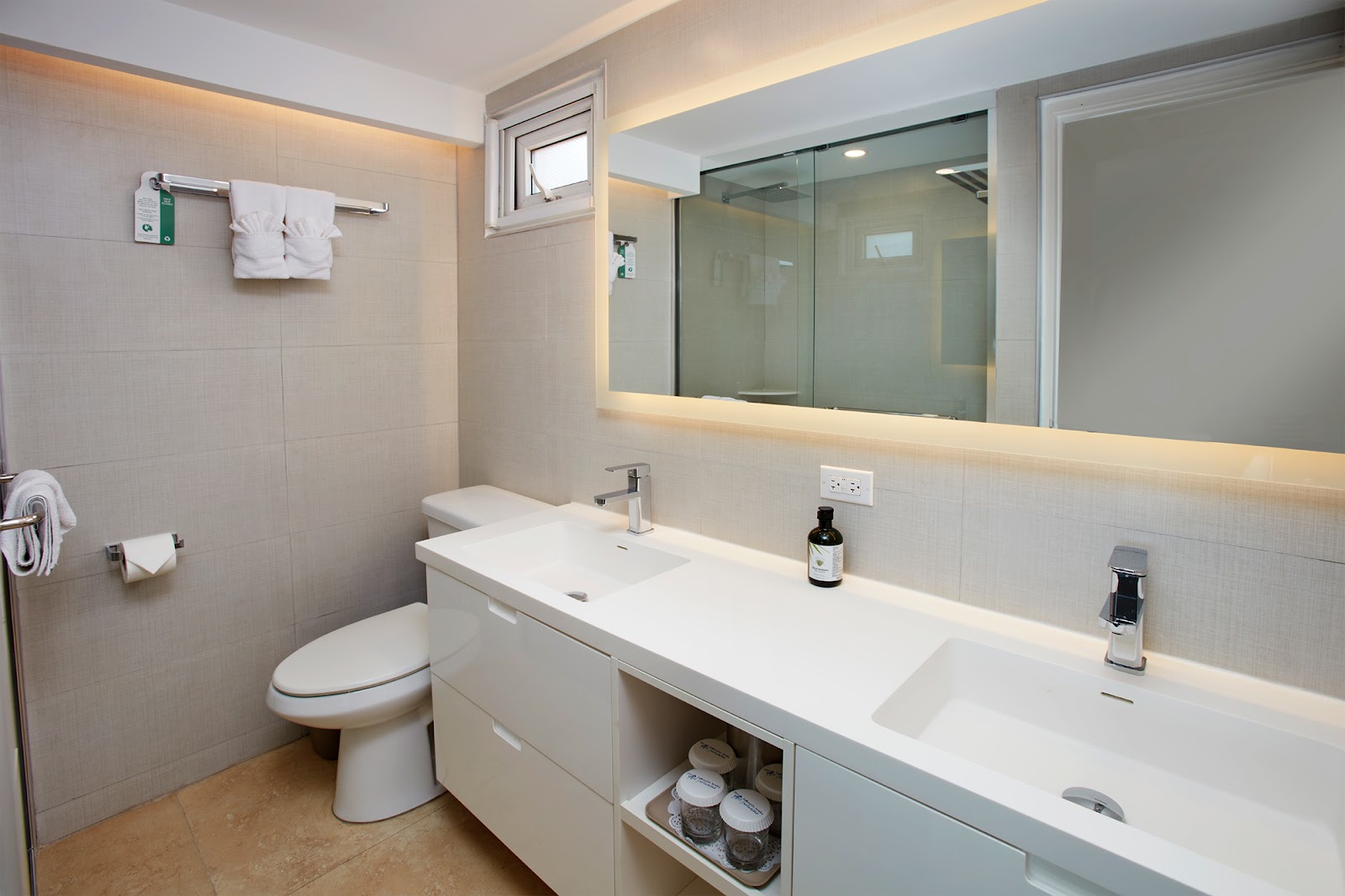 Modern bathroom with dual white sinks, lit mirror, toilet, and glass shower enclosure at Tamarijn Aruba All Inclusive Resort.