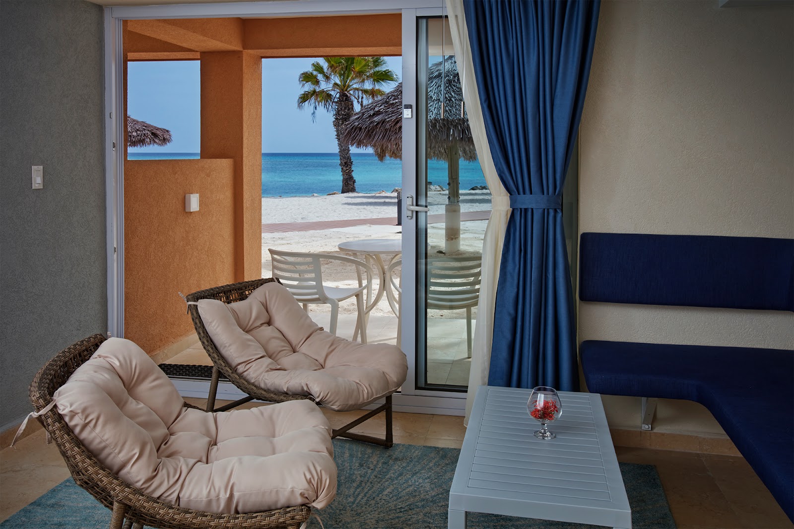 Resort room with woven chairs and ocean view through open doors to beach with palm trees and turquoise water.