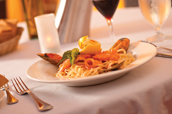Pasta with shrimp and vegetables on a white plate at a formal dining table with wine glasses.