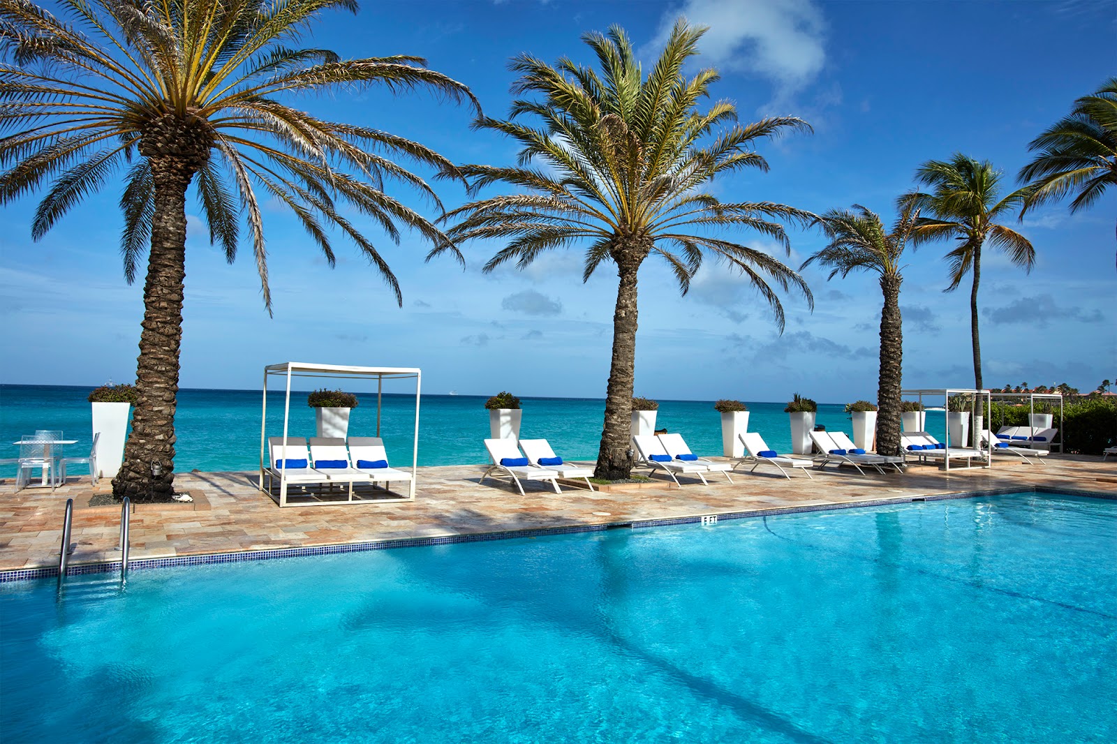 Pool with turquoise water, lounge chairs with blue cushions, white canopies, and palm trees overlooking the Caribbean Sea at Tamarijn Aruba.