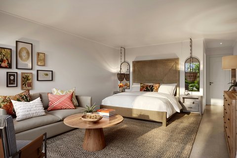 Spacious resort suite at Tamarind featuring a bed with gold headboard, seating area with gray sofa, round wooden table, and framed artwork on white walls.