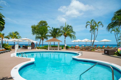 Tamarind Barbados pool area with turquoise water, white umbrellas, palm trees, and ocean view under blue sky.