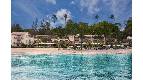 Tamarind resort buildings with cream-colored facades line a sandy beach beside turquoise water, with palm trees and tropical vegetation under a blue sky.