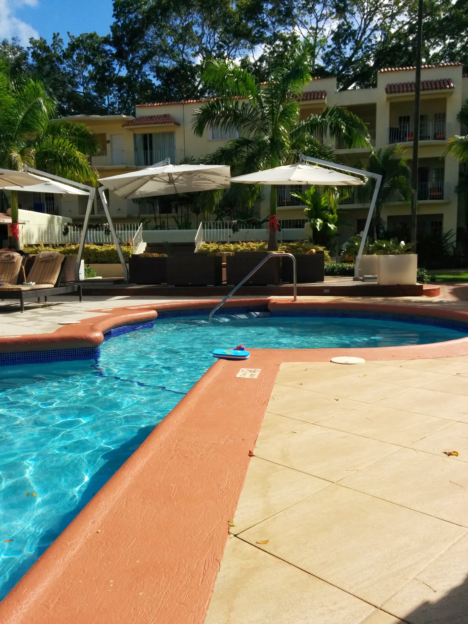 Pool area at Tamarind Barbados with clear turquoise water, terracotta deck, white cantilever umbrellas, and cream-colored resort buildings with palm trees in background.