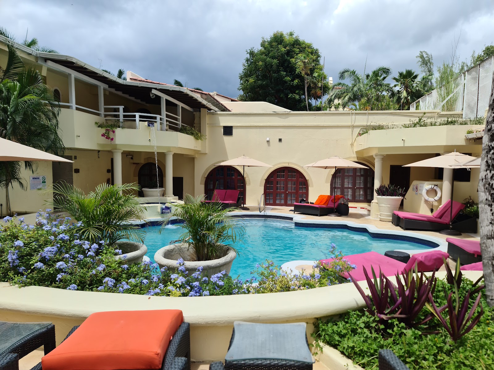 Courtyard pool at Tamarind with turquoise water, pink lounge chairs, flowering plants, and cream-colored colonial architecture under cloudy sky.