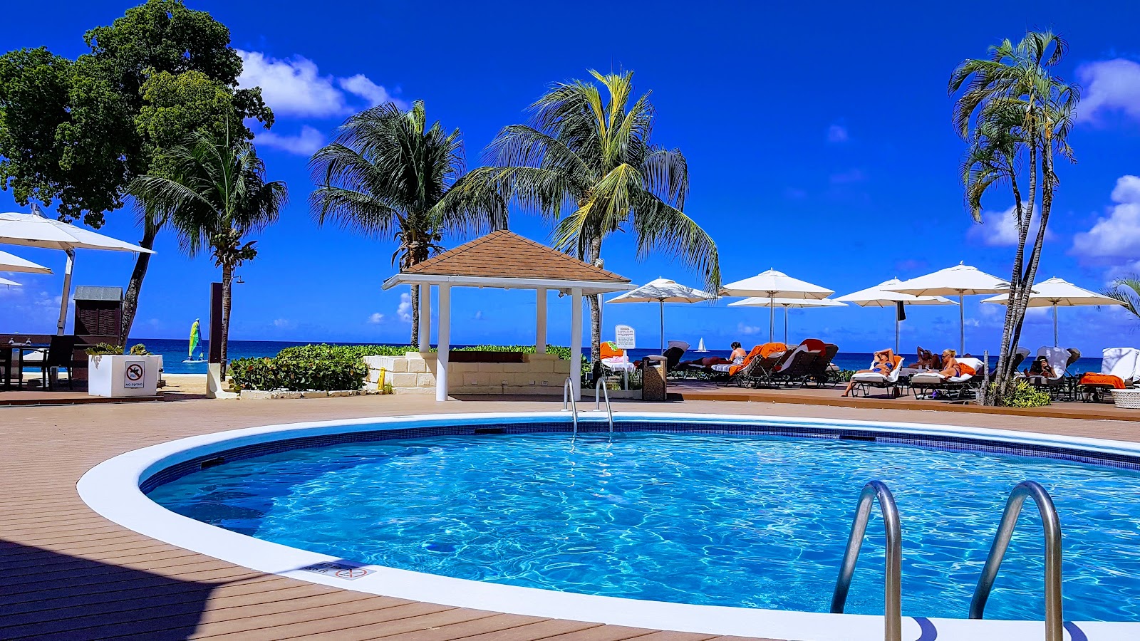 Curved pool with white edge on wooden deck at Tamarind resort, with palm trees, wooden gazebo, beach loungers under umbrellas, and ocean beyond under blue sky.