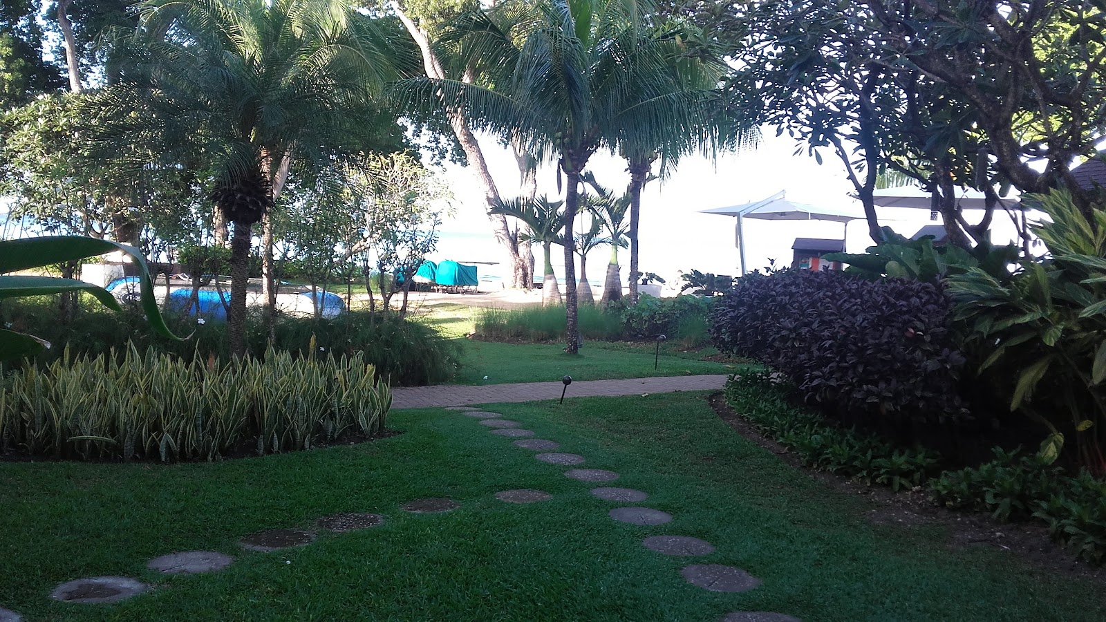 Stone pathway winds through manicured lawn flanked by palm trees and tropical plants at Tamarind Barbados, with beach and blue umbrellas visible beyond.